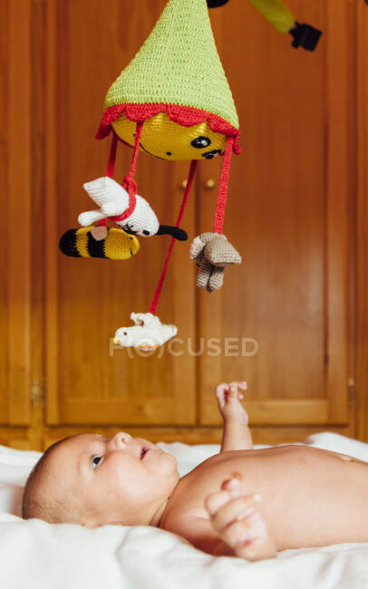 Vista lateral del alegre bebé desnudo con la boca abierta jugando con el juguete acostado en la cama divirtiéndose en casa - foto de stock