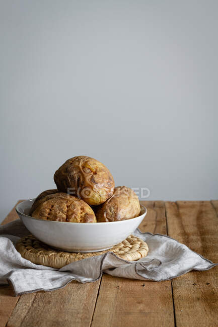 Pommes de terre marron farcies dans un bol blanc sur une serviette sur une table en bois avec un mur blanc sur le fond — Photo de stock