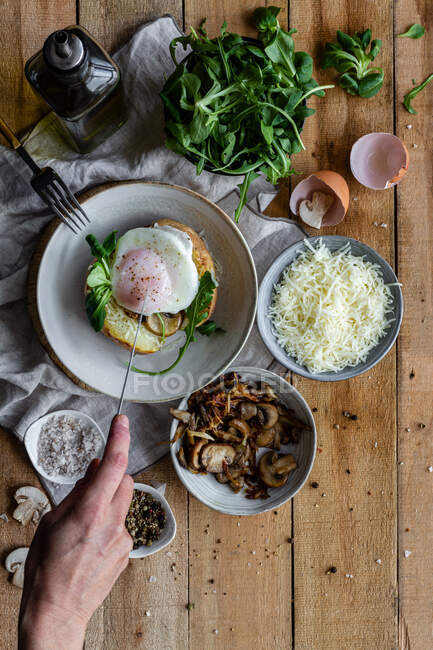 Top view of crop hand with knife cutting fried egg on potato on wooden table with fried mushrooms grated cheese and herbs — Stock Photo