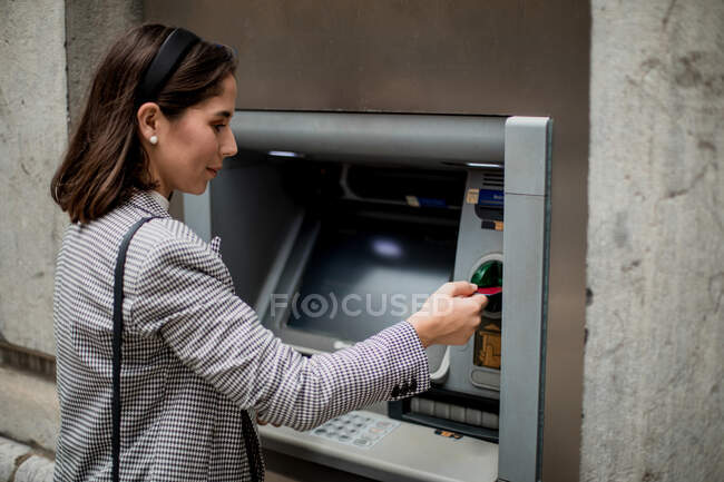 Vista lateral de una dama de pelo negro bien vestida insertando tarjeta de crédito en el cajero automático - foto de stock