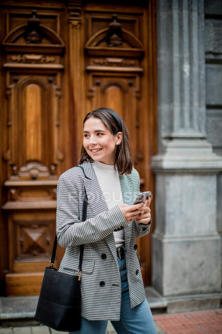 Charming young lady in stylish casual outfit smiling and looking away while surfing on mobile phone in city street — Stock Photo