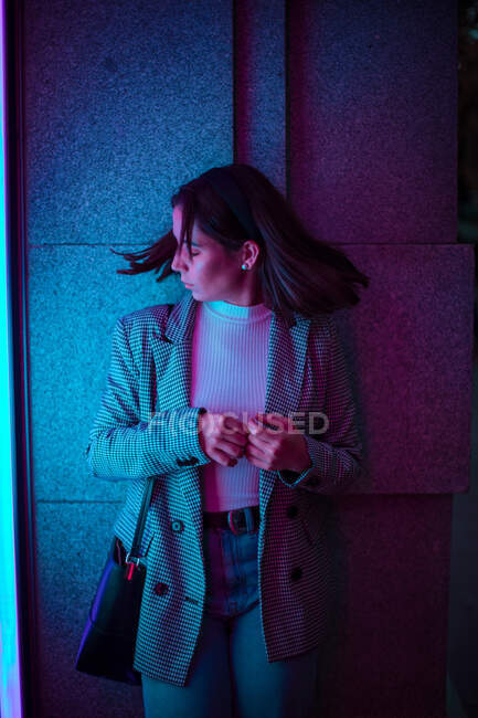 Weiblicher Teenager in lässiger Jeans und grauer Jacke steht und schüttelt den Kopf mit wehenden schwarzen Haaren im Neonlicht — Stockfoto