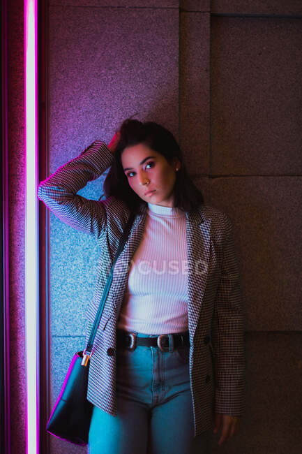 Female teenager looking at camera in casual jeans and grey jacket standing and shaking head with waving black hair in neon light — Stock Photo