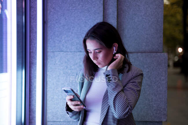Donna concentrata in elegante abbigliamento casual utilizzando auricolari wireless e telefoni cellulari in strada in città in luce al neon — Foto stock