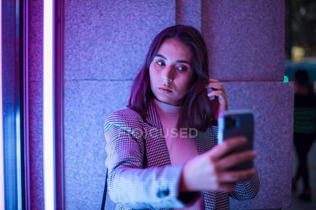 Mujer tomando fotos en teléfonos móviles tomados de la mano frente a la cámara con luz de neón - foto de stock