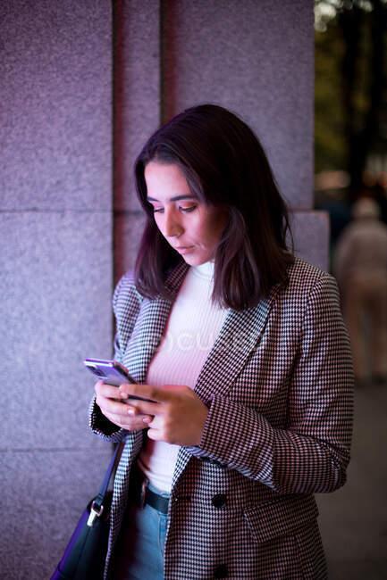 Concentrated female in stylish casual outfit using wireless earphones and mobile phones at city street in neon light — Stock Photo