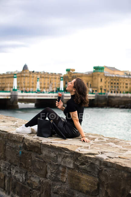 Side view of joyful black haired young lady laughing while sitting on rocked fence of quay with closed eyes holding ice cream cone with red cream — Stock Photo