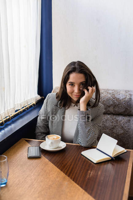 From above of cute black haired female student in stylish wear smiling and looking at camera while drinking coffee in modern cafe — Stock Photo