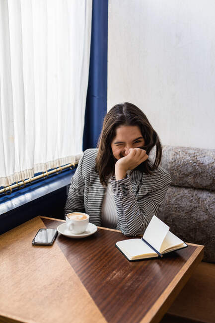 Von oben die süße schwarzhaarige Studentin in stilvoller Kleidung, die lächelt und in die Kamera schaut, während sie in einem modernen Café Kaffee trinkt — Stockfoto