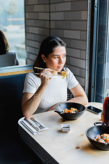 Сверху задумчивая темноволосая женщина в повседневной одежде с палочками для еды и азиатской едой с овощами, глядя в окно в кафе — стоковое фото