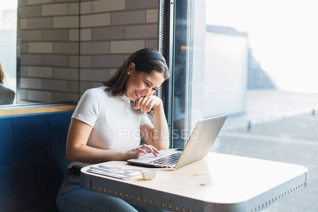Agradable dama de pelo negro en ropa casual riendo mientras se utiliza el ordenador portátil durante el almuerzo en la cafetería moderna - foto de stock