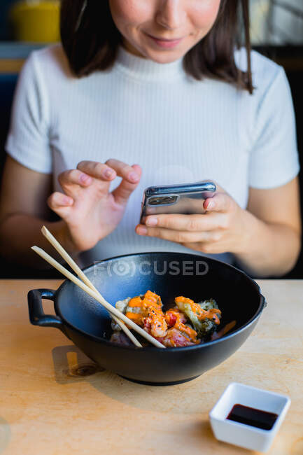 Cultivé femme aux cheveux noirs méconnaissable dans des vêtements décontractés à l'aide de téléphone mobile et de manger des aliments asiatiques avec des légumes avec baguette — Photo de stock