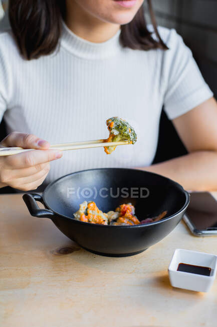 Zugeschnittene, bis zur Unkenntlichkeit nachdenkliche schwarzhaarige Frau in lässiger Kleidung, die Essstäbchen benutzt und asiatisches Essen mit Gemüse isst, während sie durch ein Fenster im Café blickt — Stockfoto