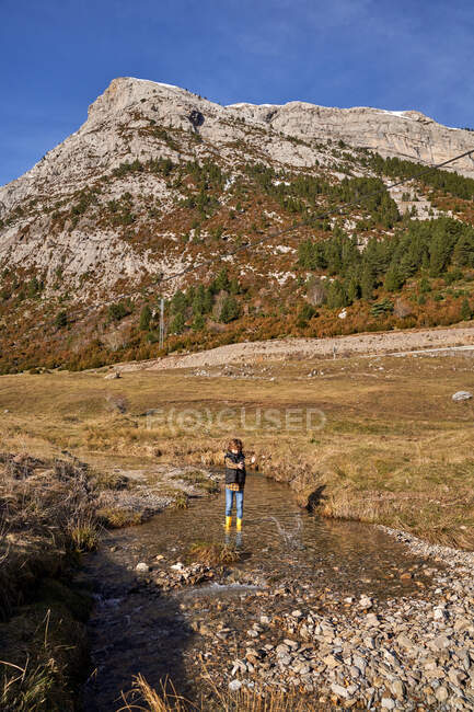 Adorable child in yellow rubber boots standing in small river at foot of stony snowy mountains in bright day — Stock Photo