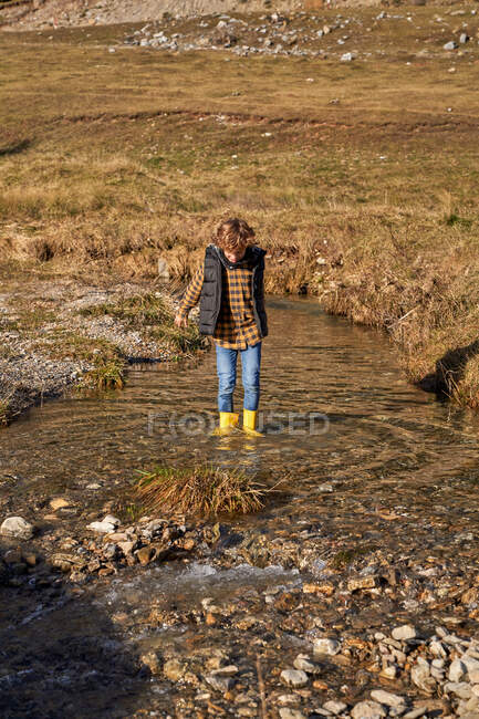 Adorable child in yellow rubber boots standing in small river at foot of stony snowy mountains in bright day — Stock Photo