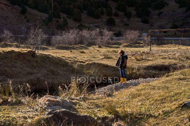 Side view of adorable kid in yellow rubber boots standing on small stony place in river at foot of stony snowy mountains with cottages in bright day — Stock Photo