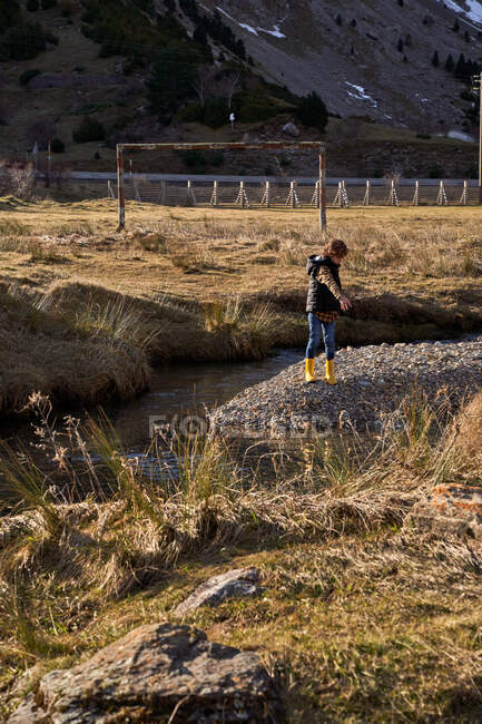 Adorable kid in yellow rubber boots standing on small stony place in river at foot of stony snowy mountains with cottages in bright day — Stock Photo