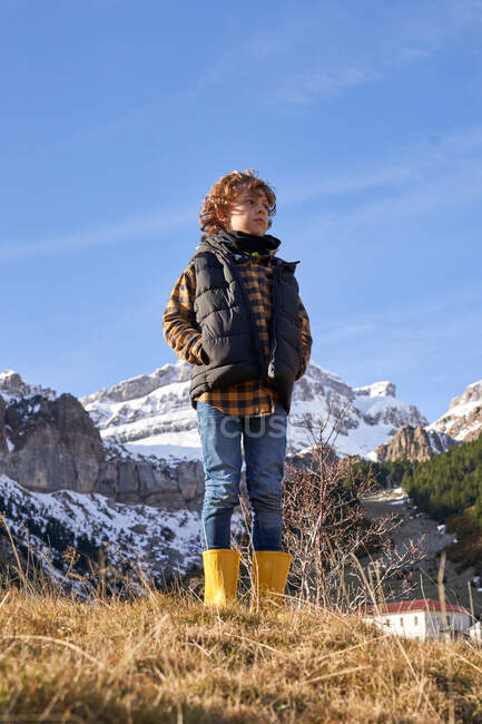 Вміст активної дитини в теплому жилеті і жовтих гумових черевиках з руками в кишені, стоячи на сухому лузі, дивлячись на пішки від засніжених гір в яскравий день — стокове фото