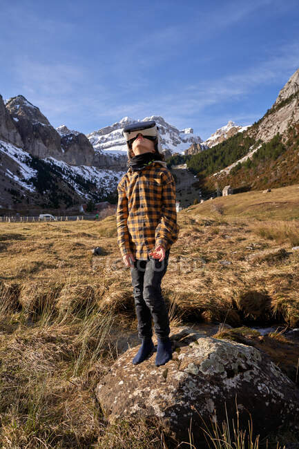 Ragazzo intelligente attivo che guarda in su in VR occhiali in piedi sulla pietra nella valle di montagna — Foto stock
