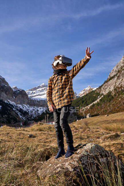 Garçon intelligent actif regardant loin dans des lunettes VR debout sur la pierre dans la vallée de la montagne — Photo de stock