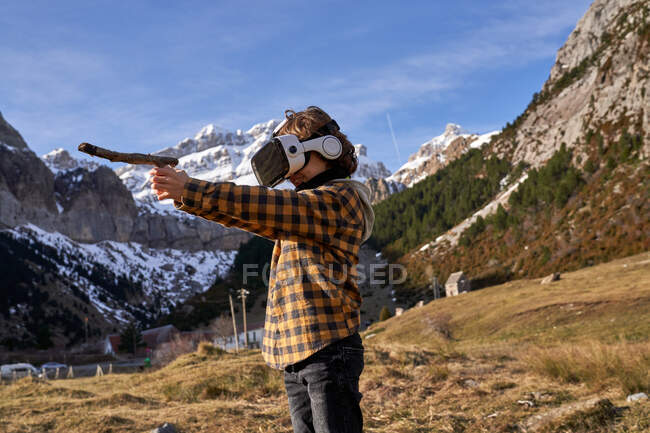 Активный умный мальчик смотрит в очки виртуальной реальности, играя с палкой, стоящей на камне в горной долине — стоковое фото