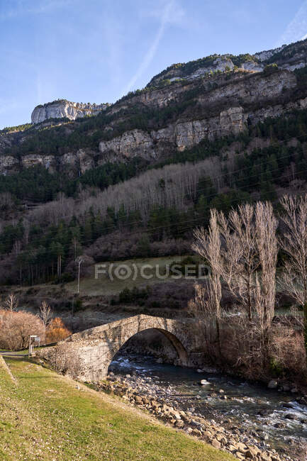 Landscape of ancient arched bridge in mountains crossing stream with dry leafless trees in bright day — Stock Photo