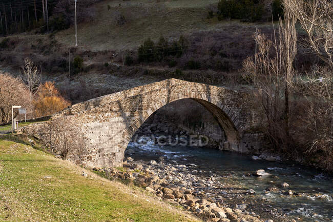 Paisaje del antiguo puente arqueado en las montañas cruzando arroyo con árboles secos sin hojas en día brillante - foto de stock