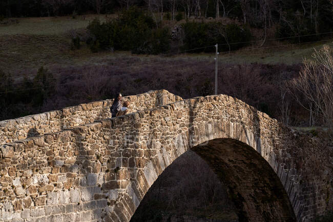 Vista posterior de la mujer viajera con el niño moviéndose a través del puente del arco antiguo al valle del bosque en día brillante - foto de stock