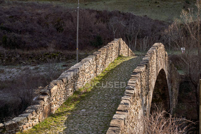 Paisagem da ponte arqueada antiga em montanhas que cruzam o córrego com árvores sem folhas secas em dia brilhante — Fotografia de Stock