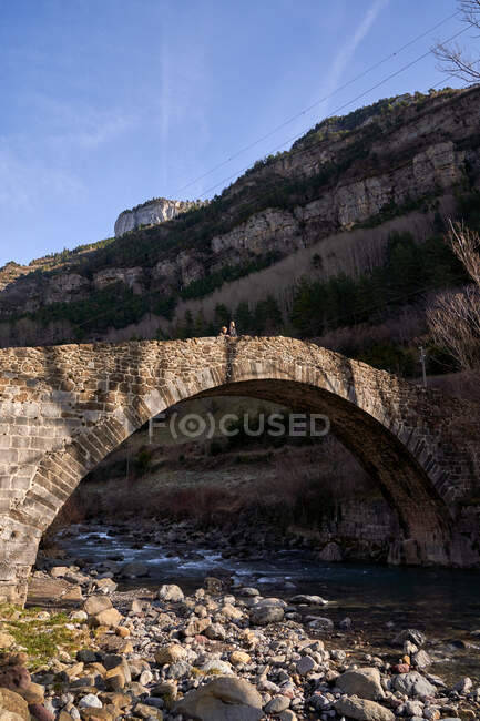 Donna e bambino apprezzano il paesaggio dell'antico ponte ad arco in montagne che attraversano il ruscello con alberi senza foglie secchi nella giornata luminosa — Foto stock
