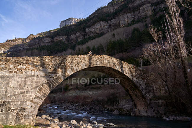 Mujer y niño apreciando el paisaje del antiguo puente arqueado en las montañas cruzando arroyo con árboles secos sin hojas en día brillante - foto de stock