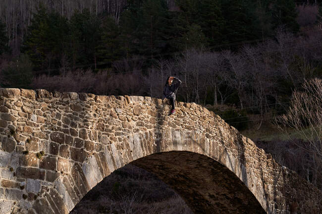 Weit weg blickende Frau sitzt bei hellem Tag auf uralter Bogenbrücke ins Waldtal — Stockfoto