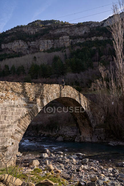 Traveling woman looking far away sitting on ancient arch bridge to forest valley in bright day — Stock Photo