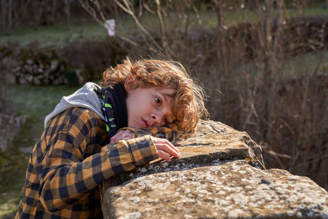 Side view of curly pensive tender child resting lying on hand on stone fence in forest valley looking away — Stock Photo