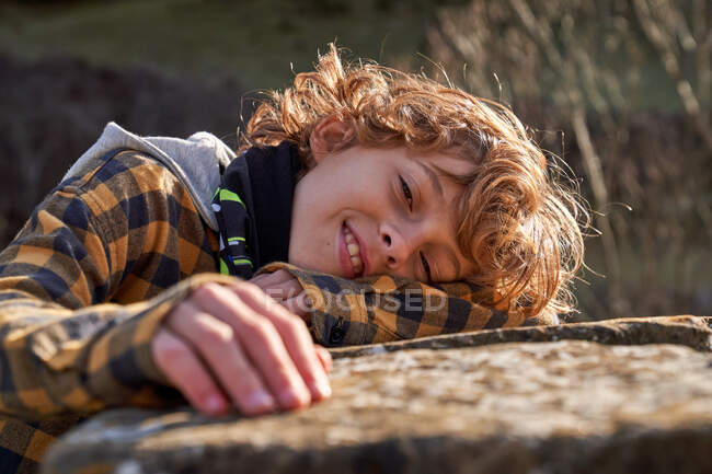 Side view of curly pensive tender child resting lying on hand on stone fence in forest valley looking at camera — Stock Photo