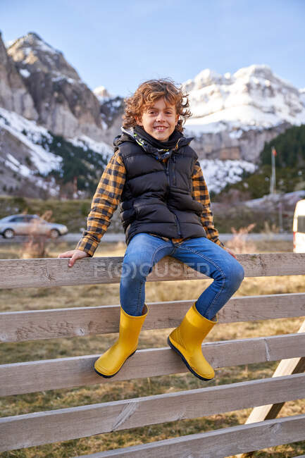 Adorable enfant joyeux en gilet chaud et bottes en caoutchouc jaune assis sur une vieille clôture en bois dans le village au pied de montagnes enneigées et regardant la caméra — Photo de stock