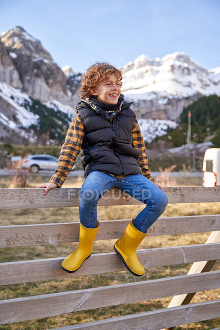 Adorable joyful kid in warm vest and yellow rubber boots sitting on old wooden fence in village at foot of snowy mountains and looking away — Stock Photo