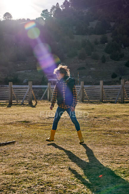 Fröhlich-enthusiastisches Kind in warmer Weste und gelben Gummistiefeln spielt bei sonnigem Wetter im leeren Hof eines Dorfes am Fuße der Berge — Stockfoto