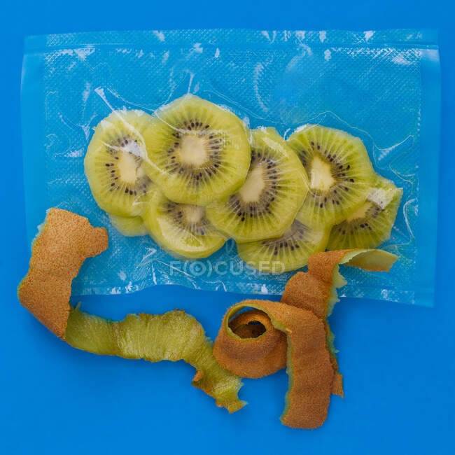 Vista dall'alto di kiwi pelati maturi in sacchetto di plastica sottovuoto e buccia marrone su sfondo blu — Foto stock