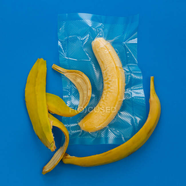 Top view of ripe yellow peeled banana in vacuum plastic bag and banana peel on blue background — Stock Photo