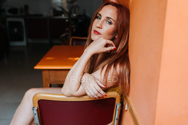 Side view from above of redhead content woman laughing and looking at camera while resting on chair in modern kitchen — Stock Photo