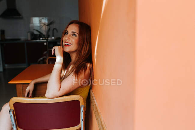 Side view from above of redhead content woman laughing and looking away while resting on chair in modern kitchen — Stock Photo