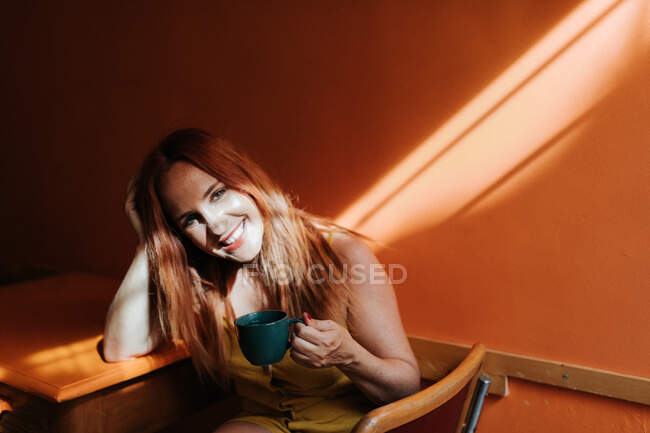 Vista laterale dall'alto della donna dai contenuti rossi che ride e guarda la fotocamera mentre riposa sulla sedia nella cucina moderna — Foto stock