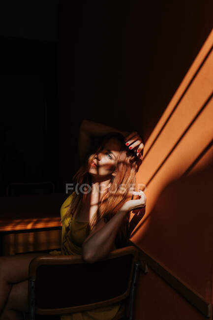 Vista lateral do modelo pensativo no vestido amarelo elegante com maquiagem sentada na cadeira e olhando para longe — Fotografia de Stock