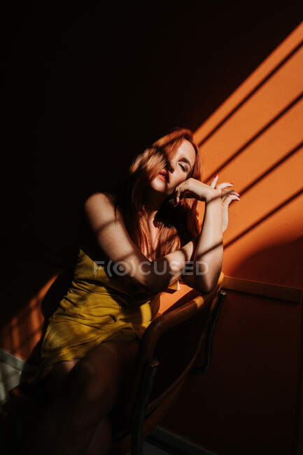 Sensual modelo femenino pelirroja reflexivo en elegante vestido amarillo con maquillaje sentado con las piernas cruzadas en la silla bajo el haz de sol en habitación oscura - foto de stock