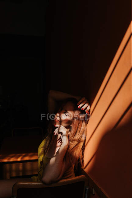 Вид сбоку чувственной рыжеволосой модели в стильном желтом платье с макияжем, сидящей на стуле с закрытыми глазами под лучами солнца в темной комнате — стоковое фото