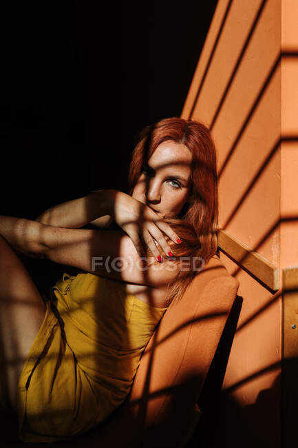 Sinnliches Model in stylischem gelben Kleid mit Make-up sitzt auf Stuhl und schaut in die Kamera unter dem Sonnenstrahl im dunklen Raum — Stockfoto