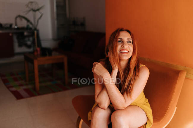 Mujer pelirroja alegre mirando hacia otro lado mientras descansa en la silla en casa - foto de stock