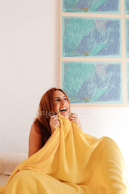 High angle of playful redhead woman sitting and covering half of face with yellow blanket while looking away on bed — Stock Photo