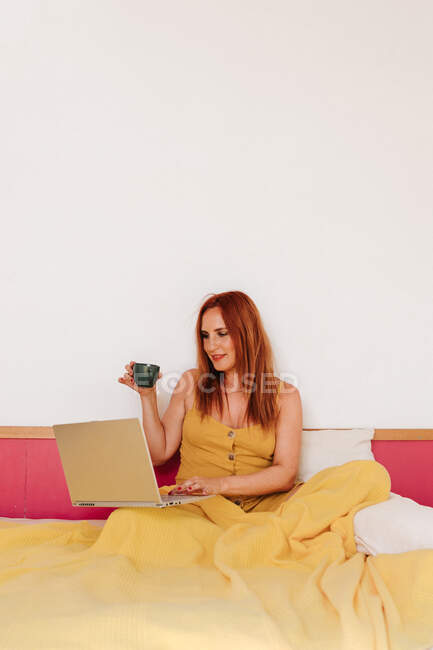 Rothaarige Freiberuflerin in gelbem Kleid arbeitet mit Laptop, während sie Kaffee trinkt und auf dem Bett liegt — Stockfoto